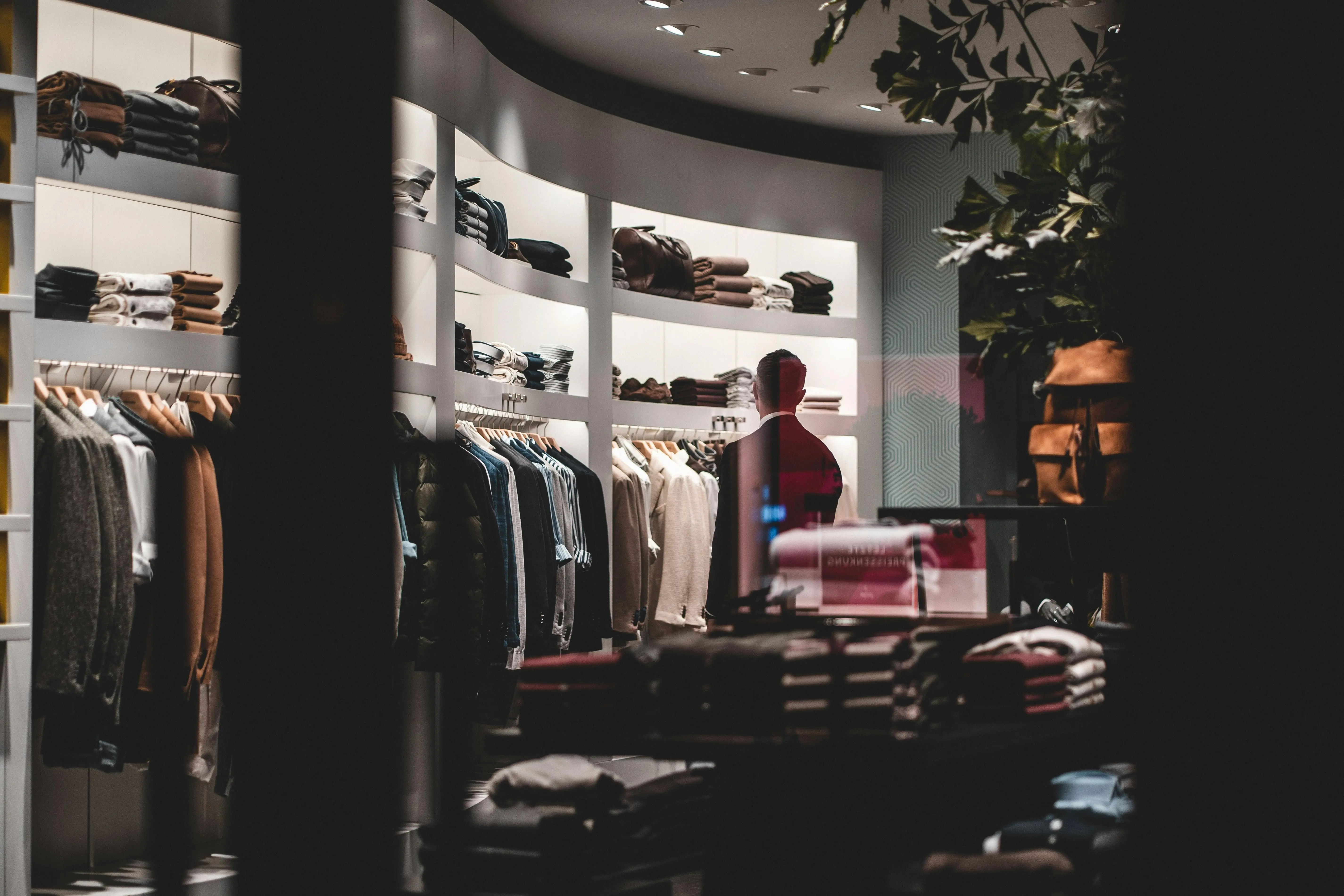 Man viewed through a store window looking at shelves and racks of folded clothes and hanging jackets in a dimly lit clothing shop.