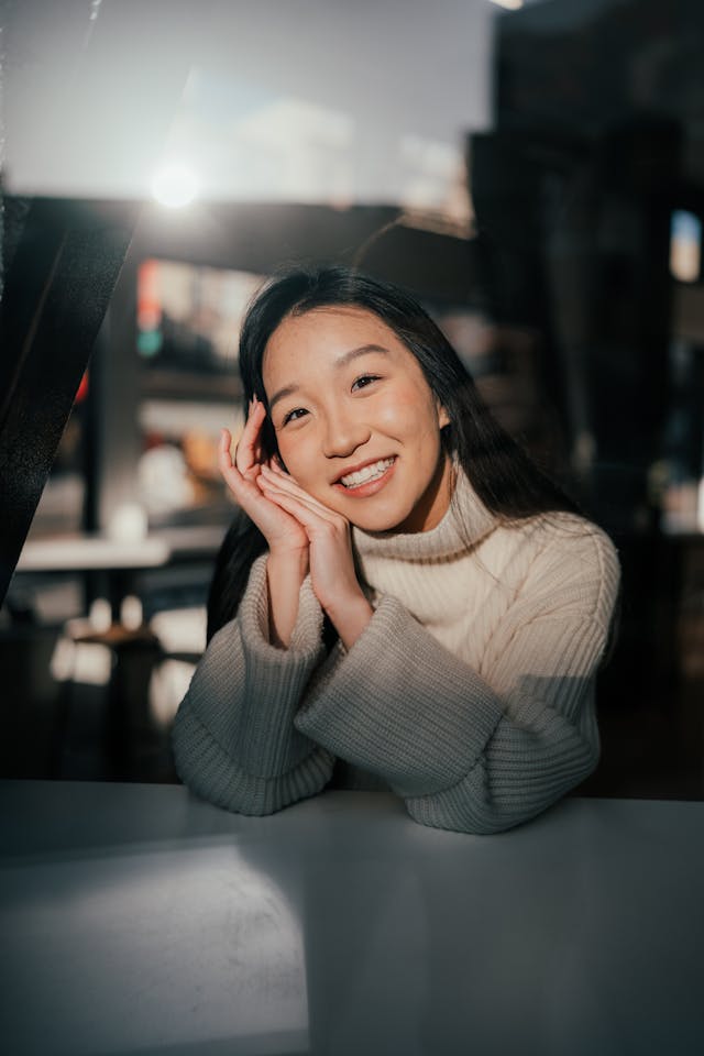 Smiling young woman in a cream turtleneck sweater sitting at a table with her hands gently touching her face, lit by soft natural light through a window.