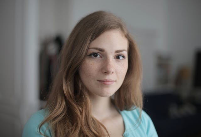 Young woman with light brown wavy hair and freckles wearing a light blue top, looking directly at the camera with a neutral expression.